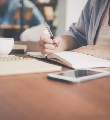 woman writing on a notebook beside teacup and tablet computer